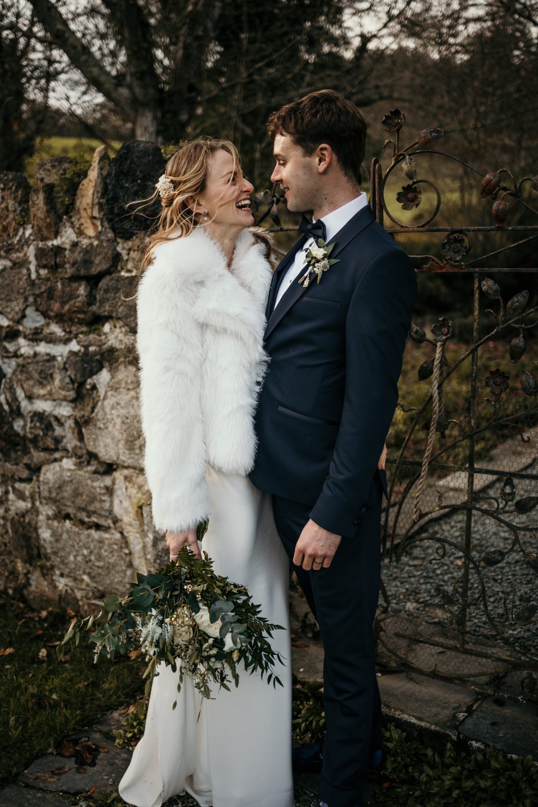 bride and groom pose in the grounds of the great barn devon during their winter wedding reception. the bride wears fur jacket