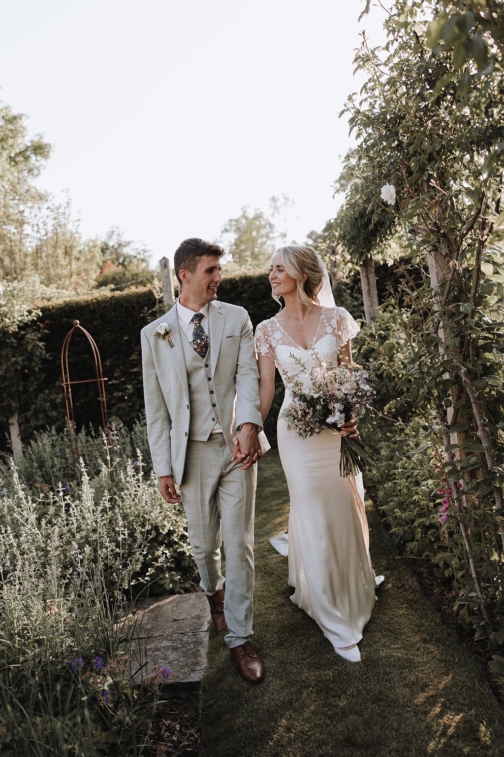 bride and groom walking through gardens at the great barn devon, the bride carries her bouquet 